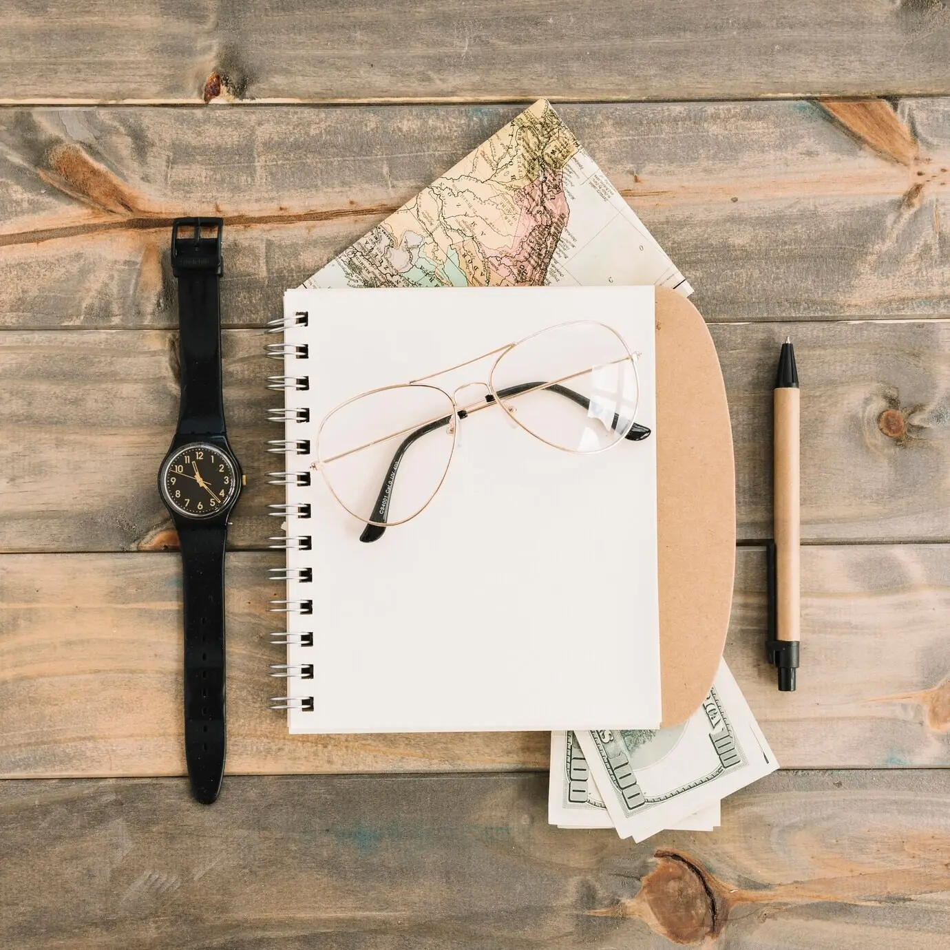 Overhead view of eyeglasses atop a spiral notebook, with currency, a map, a wristwatch, and a pen on a wooden plank background.